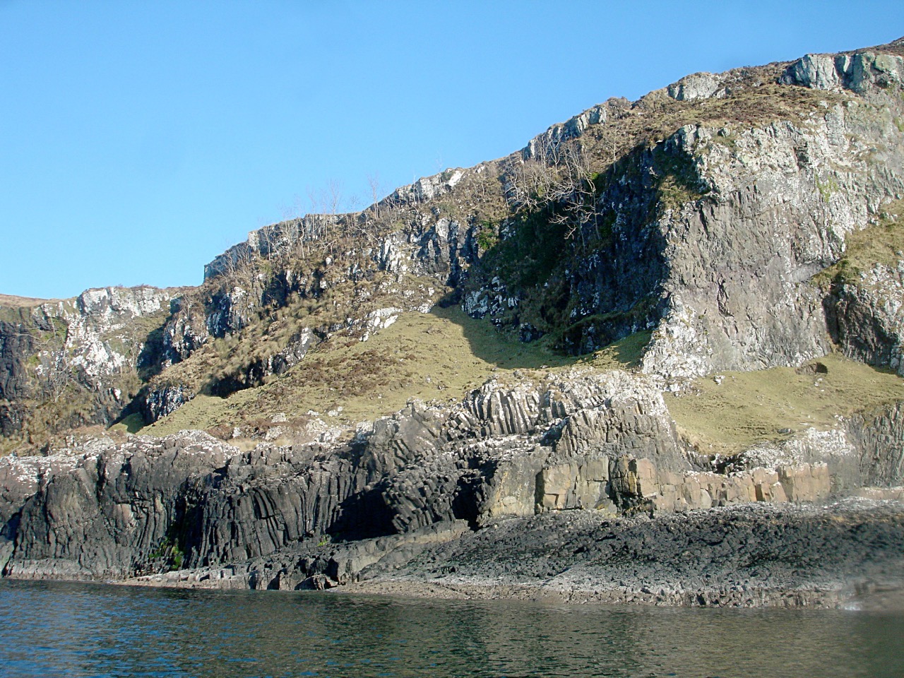Dark charcoal-grey columnar basalt cliffs meeting the sea on the Isle of Kerrera, Scotland.