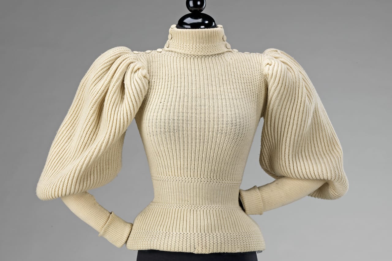 A cream ribbed wool sweater with a high buttoned collar and full gathered sleeves, shown on a dress form against a grey backdrop.