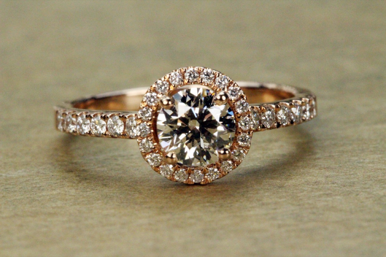 A rose gold ring with a round diamond surrounded by a halo of smaller diamonds, on a warm neutral surface.