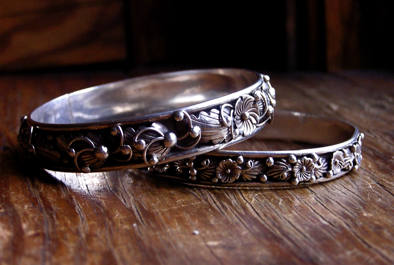 Two ornately floral-engraved silver bangle bracelets resting on a dark wood surface.