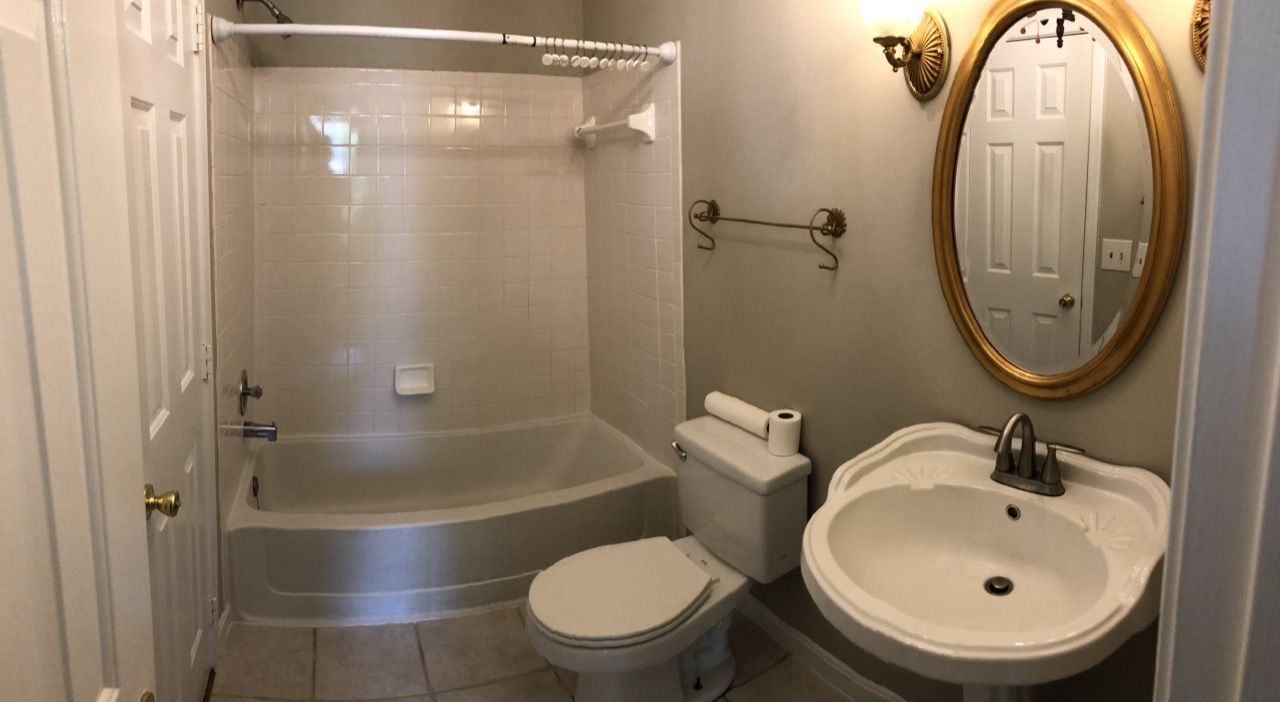 A panoramic view of a bathroom with a bathtub, toilet, warm-toned vanity and an oval mirror.