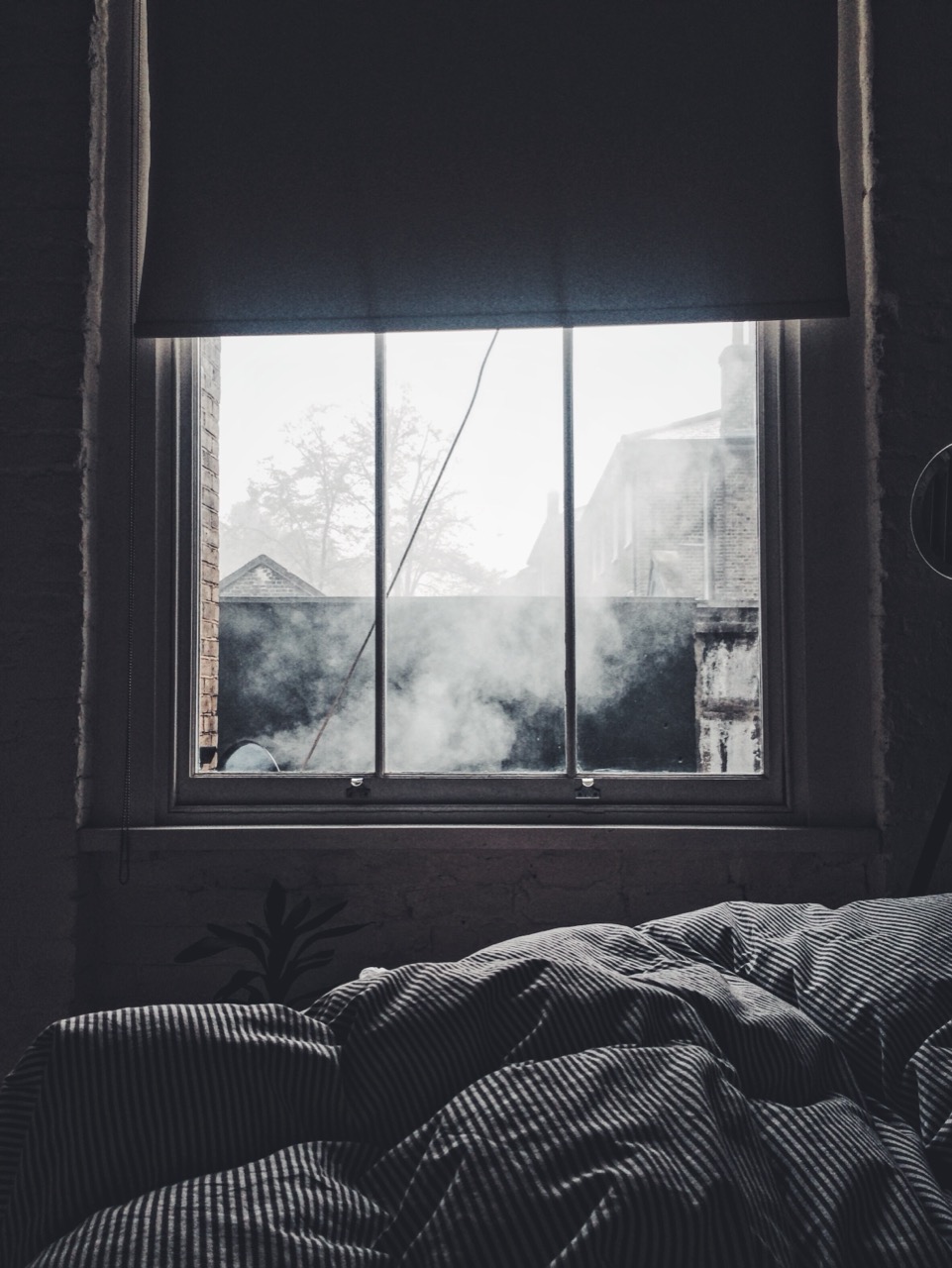 A window above an unmade bed, with soft diffuse daylight and a brick building visible outside.