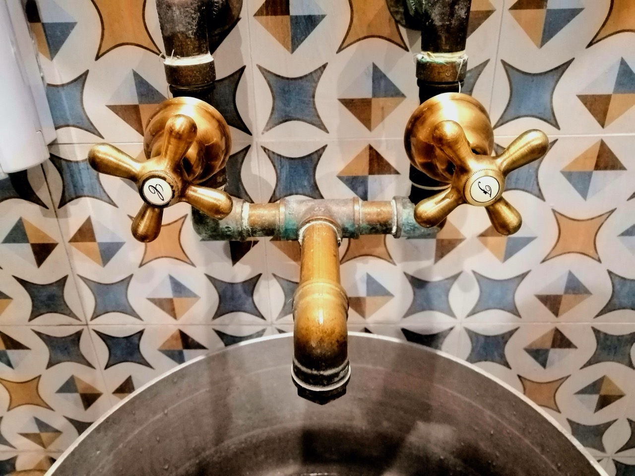 An unbranded warm-toned brass cross-handle tap against a patterned tiled wall.