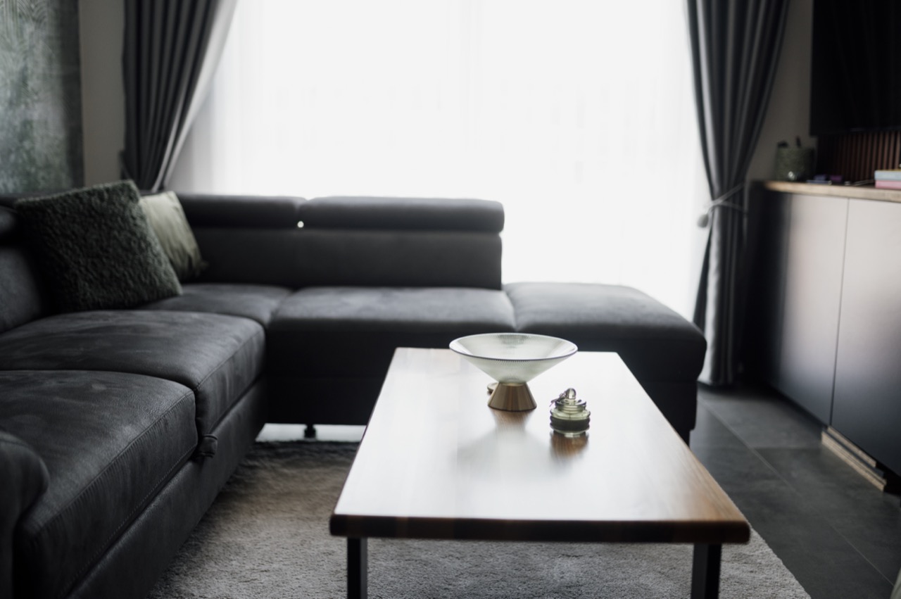A dark grey sectional sofa in a simply furnished living room lit by a bright window.